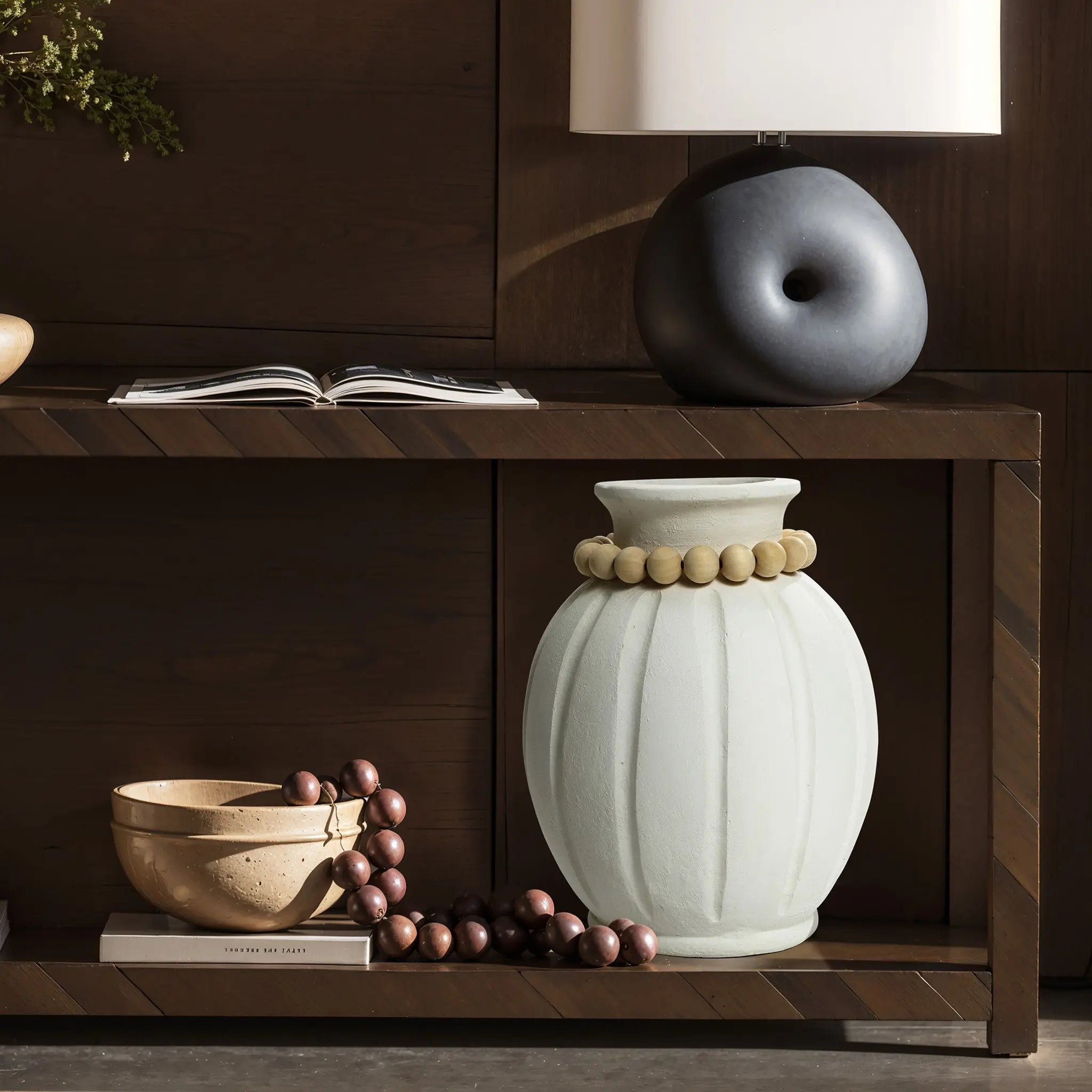A wooden shelf showcases a modern round vase adorned with beads, alongside a bowl of round objects, a black abstract sculpture, a closed book, and a minimalist lamp. The arrangement embodies minimalist design while adding elegance to the space.