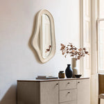 An oval, irregularly shaped wall mirror with a natural finish wood frame hangs above a beige sideboard adorned with books and vases. Sunlight streams through the window, enhancing the modern home decor.