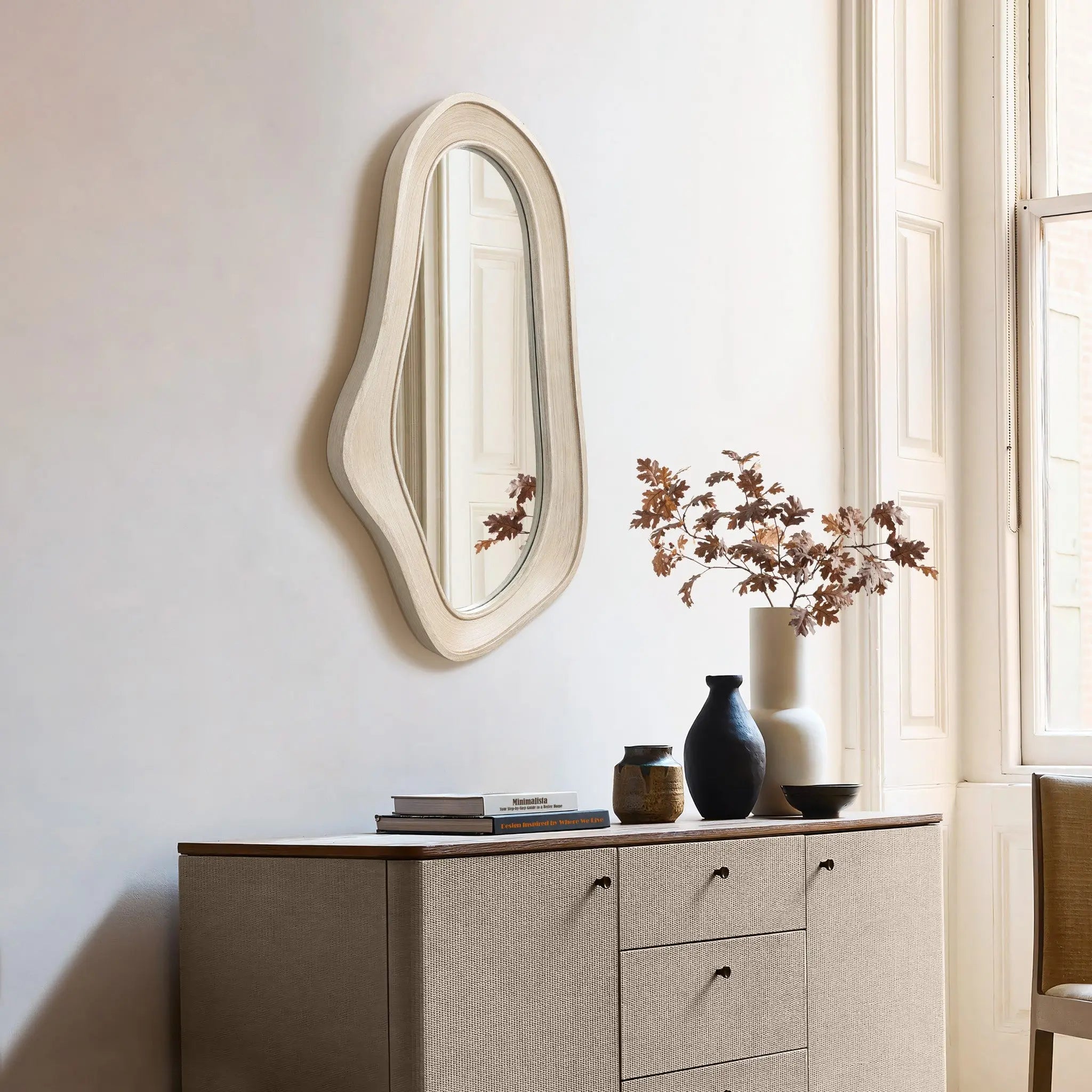 An oval, irregularly shaped wall mirror with a natural finish wood frame hangs above a beige sideboard adorned with books and vases. Sunlight streams through the window, enhancing the modern home decor.
