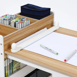 The LuxenHome Children Play Table with Bookcase and 2 Chairs Set includes a roll of paper, two markers, and a multi-compartment wooden organizer for colored markers. A blue pencil case sits on it, while the shelf below stores books—ideal for creative exploration.