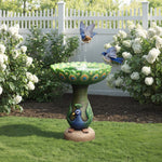 The exquisite Peacock Resin Birdbath graces the garden, surrounded by white flowers. Two birds dance above, one joyfully splashing in the basin, adding charm to the outdoor space.