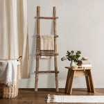 Rustic charm meets modern functionality with this solid wood blanket ladder, draped with a towel beside a small wooden table adorned with stacked towels, a plant vase, and decor, all nestled next to a wicker basket and curtain.