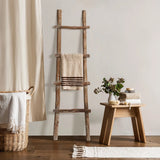 Rustic charm meets modern functionality with this solid wood blanket ladder, draped with a towel beside a small wooden table adorned with stacked towels, a plant vase, and decor, all nestled next to a wicker basket and curtain.