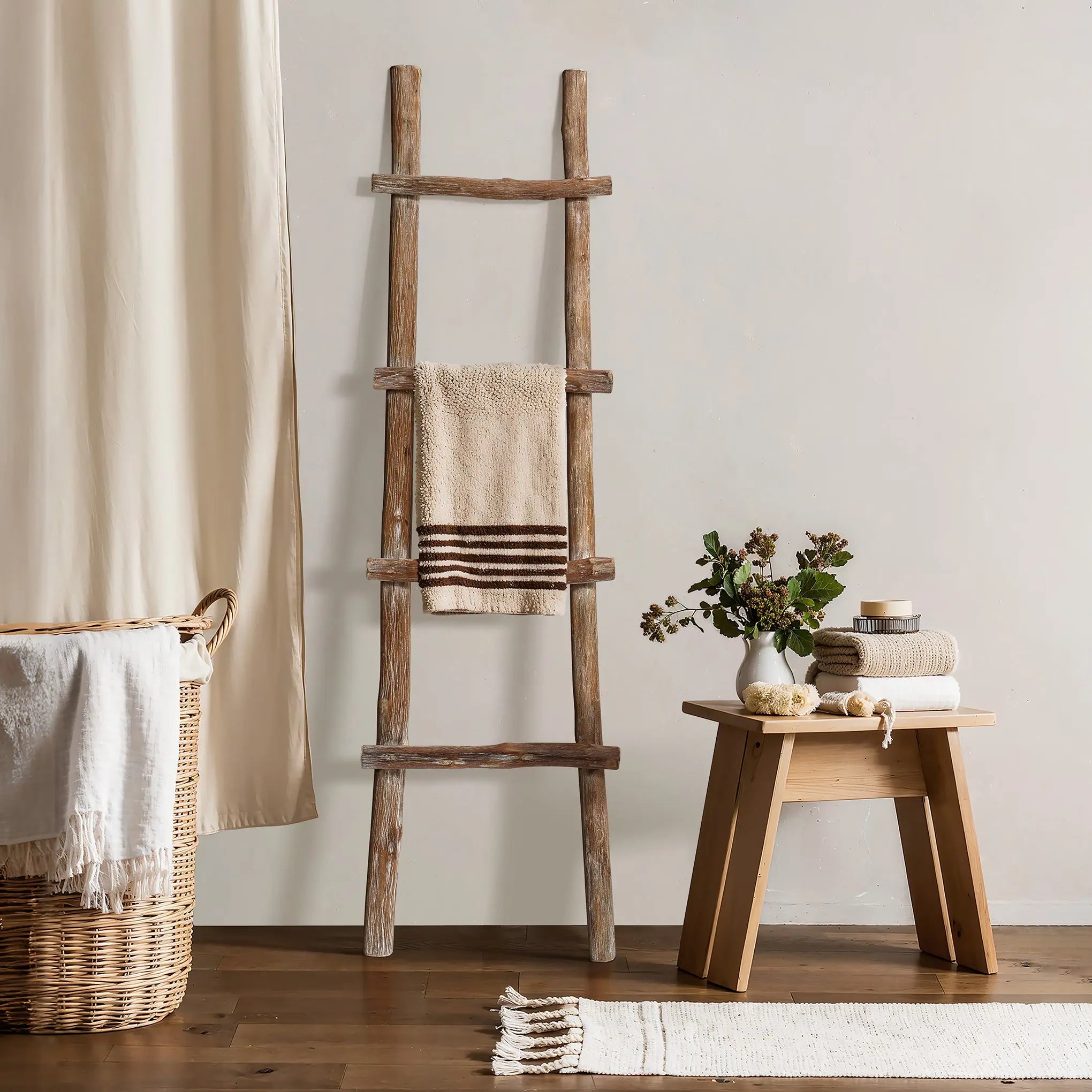 Rustic charm meets modern functionality with this solid wood blanket ladder, draped with a towel beside a small wooden table adorned with stacked towels, a plant vase, and decor, all nestled next to a wicker basket and curtain.