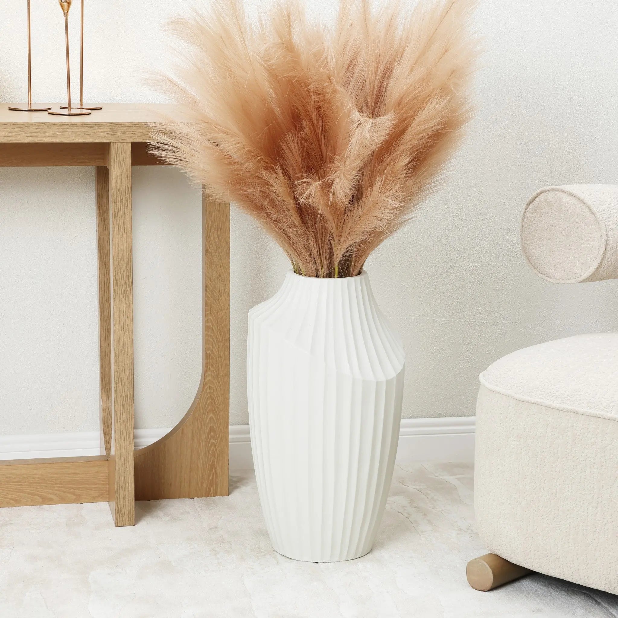 A white fluted vase, reminiscent of a modern abstract sculpture, holds pampas grass atop a light carpet beside a wooden table and beige armchair in this minimalist room.