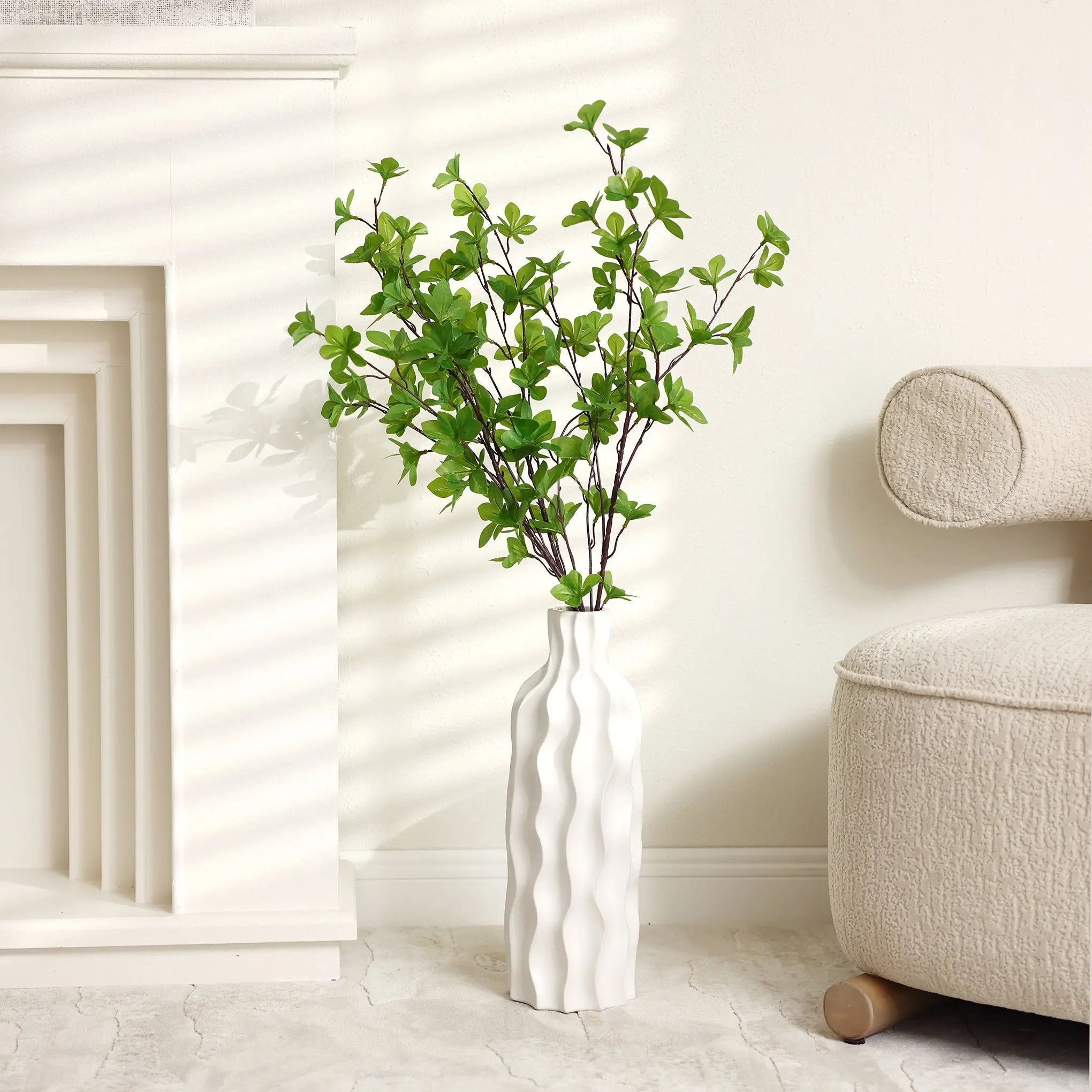 A versatile piece, this elegant ceramic vase with green leafy branches stands on a light-colored floor next to a beige sofa in a sunlit room.