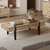 A modern farmhouse coffee table with an oak finish wood top and black legs, holding a tray with a vase of flowers and books, sits on a textured rug in a modern living room with wood flooring and light-colored furniture.