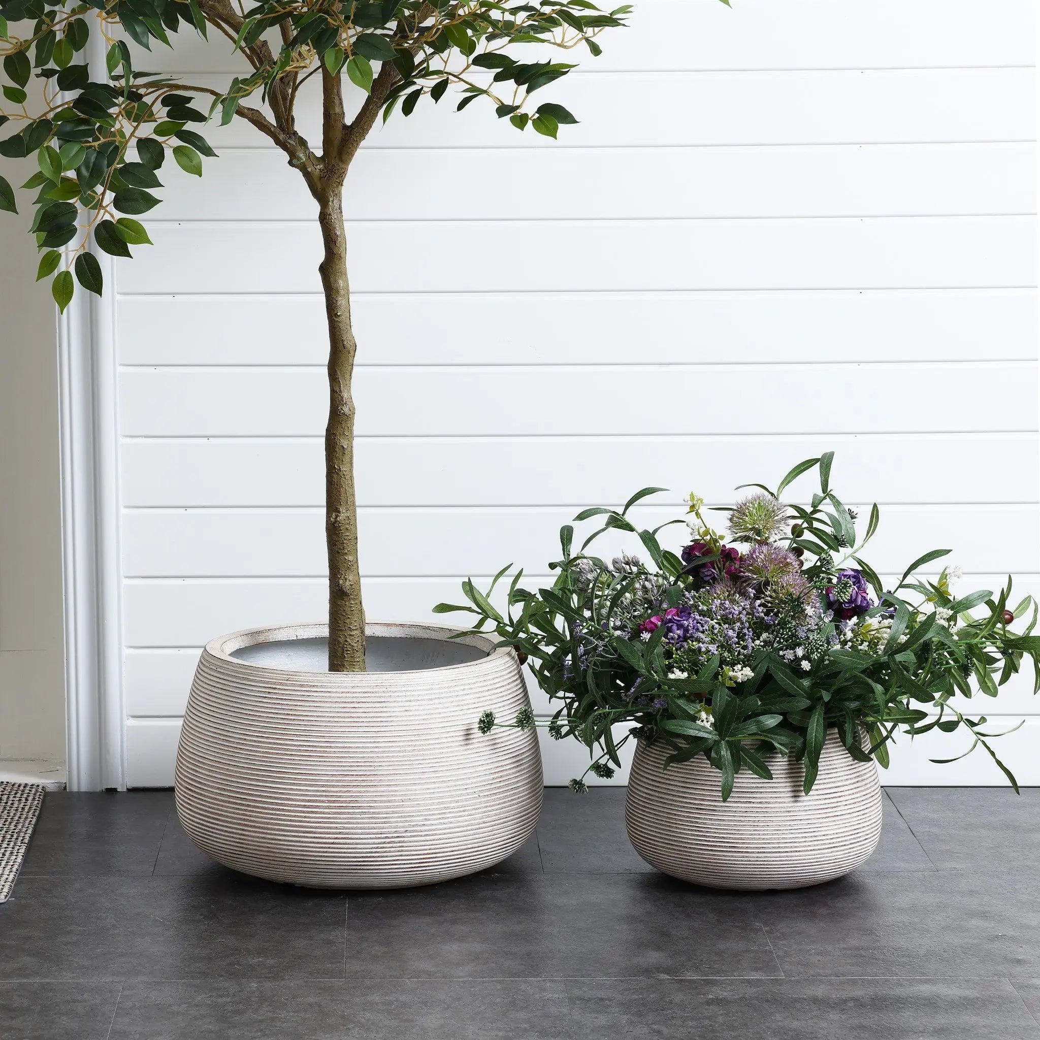 Two large speckled off-white planters sit on a gray tile floor in front of a white paneled wall. The left holds a small tree, while the right bursts with colorful flowers and greenery, embodying a minimalist design with their high-quality MgO construction.