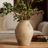 The 13.2 h Rustic beige ribbed terracotta urn vase by LuxenHome holds green leafy branches with small brown flowers on a wooden table in a cozy living room, where a beige sofa and stacked books enhance the charm.