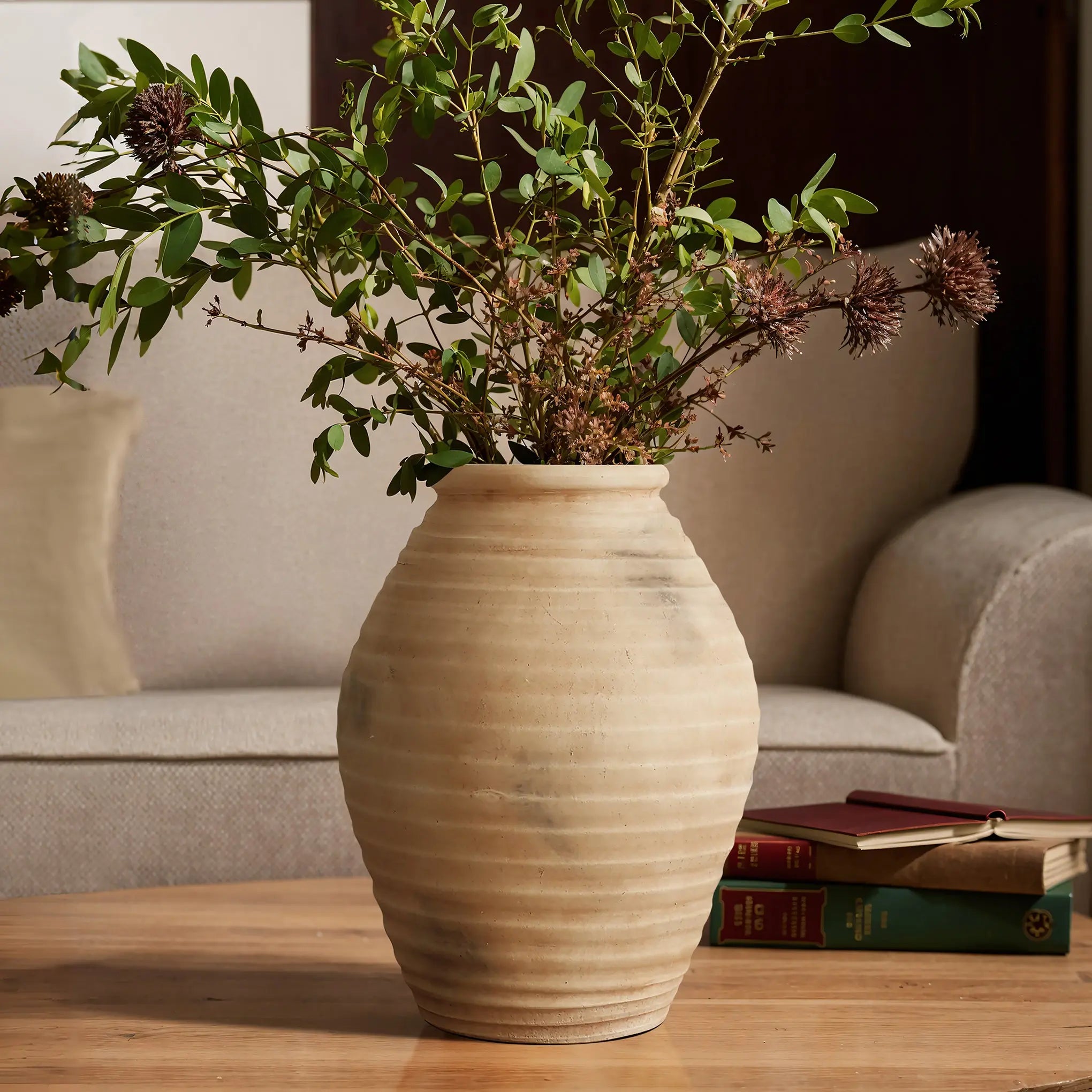 The 13.2 h Rustic beige ribbed terracotta urn vase by LuxenHome holds green leafy branches with small brown flowers on a wooden table in a cozy living room, where a beige sofa and stacked books enhance the charm.