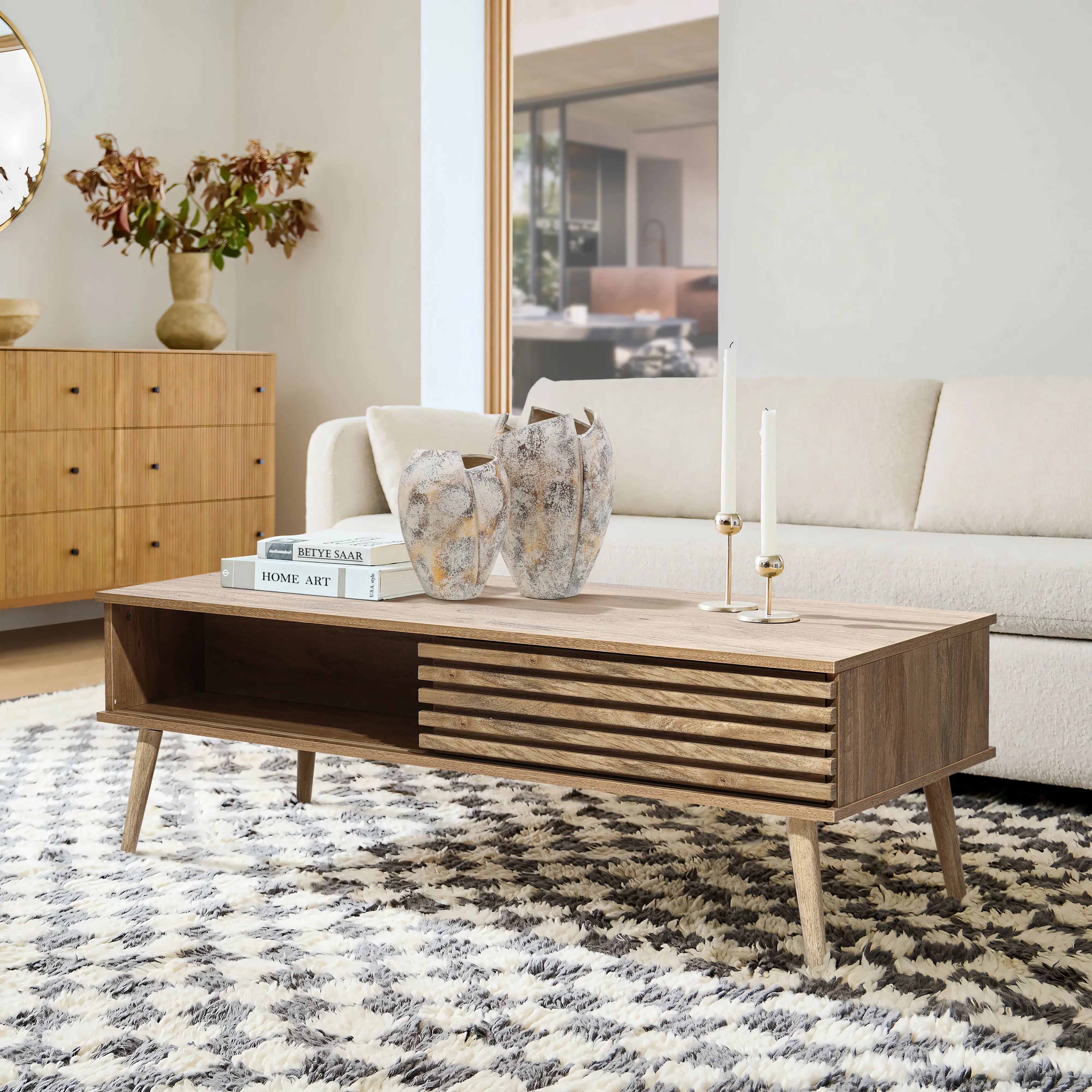 Modern living room with a light wood coffee table featuring dual-side storage, flanked by two vases and candlesticks. A beige sofa and patterned rug complement the decor, while a large window provides natural light.