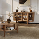 A cozy living room highlights a wooden coffee table with a vase and open book, paired with the LuxenHome Miko sideboard server cabinet featuring solid wood legs filled with decor items. A modern painting, botanical arrangement, and textured rug enhance the warmth and style of the neutral-toned space.