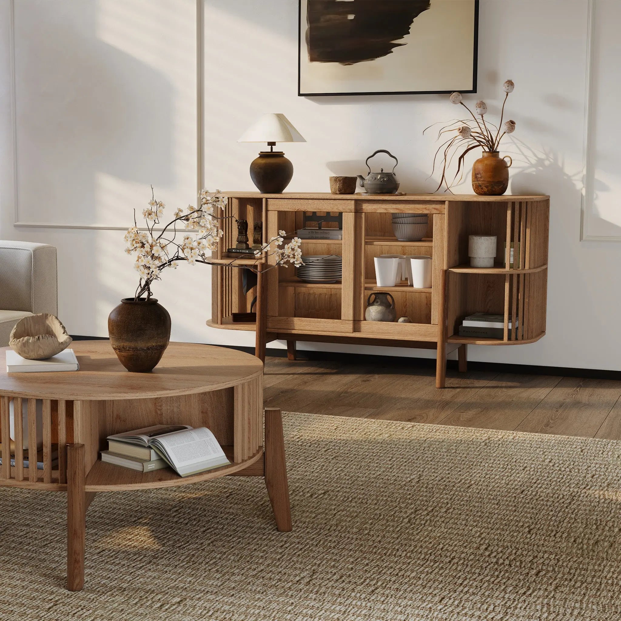 A cozy living room highlights a wooden coffee table with a vase and open book, paired with the LuxenHome Miko sideboard server cabinet featuring solid wood legs filled with decor items. A modern painting, botanical arrangement, and textured rug enhance the warmth and style of the neutral-toned space.