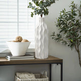 An elegant design unfolds as the white wavy ceramic vase with greenery graces the table, accompanied by a bowl of decorative balls, books, and a basket. Light filters softly through the blinds, highlighting this versatile piece in its tranquil setting.