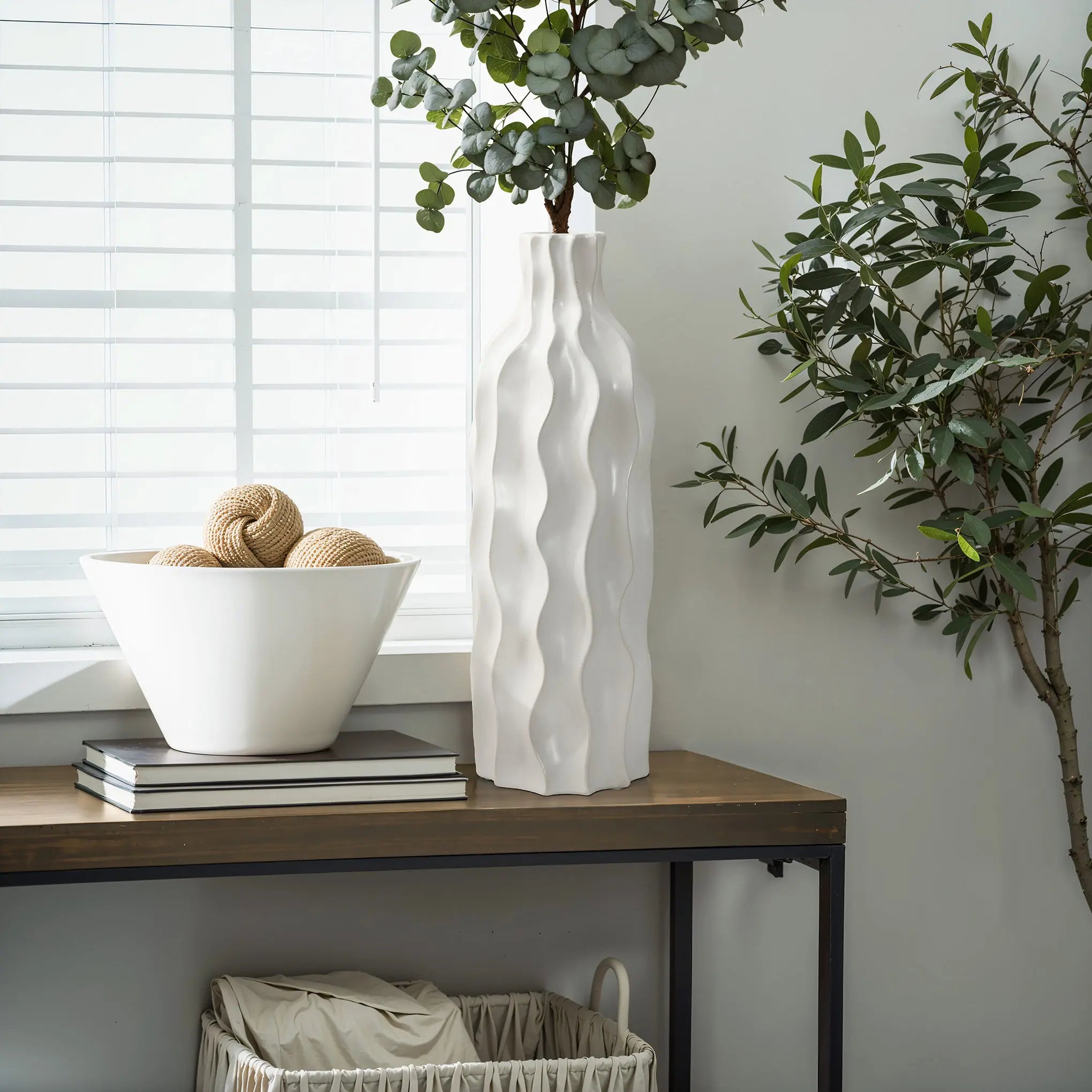 An elegant design unfolds as the white wavy ceramic vase with greenery graces the table, accompanied by a bowl of decorative balls, books, and a basket. Light filters softly through the blinds, highlighting this versatile piece in its tranquil setting.
