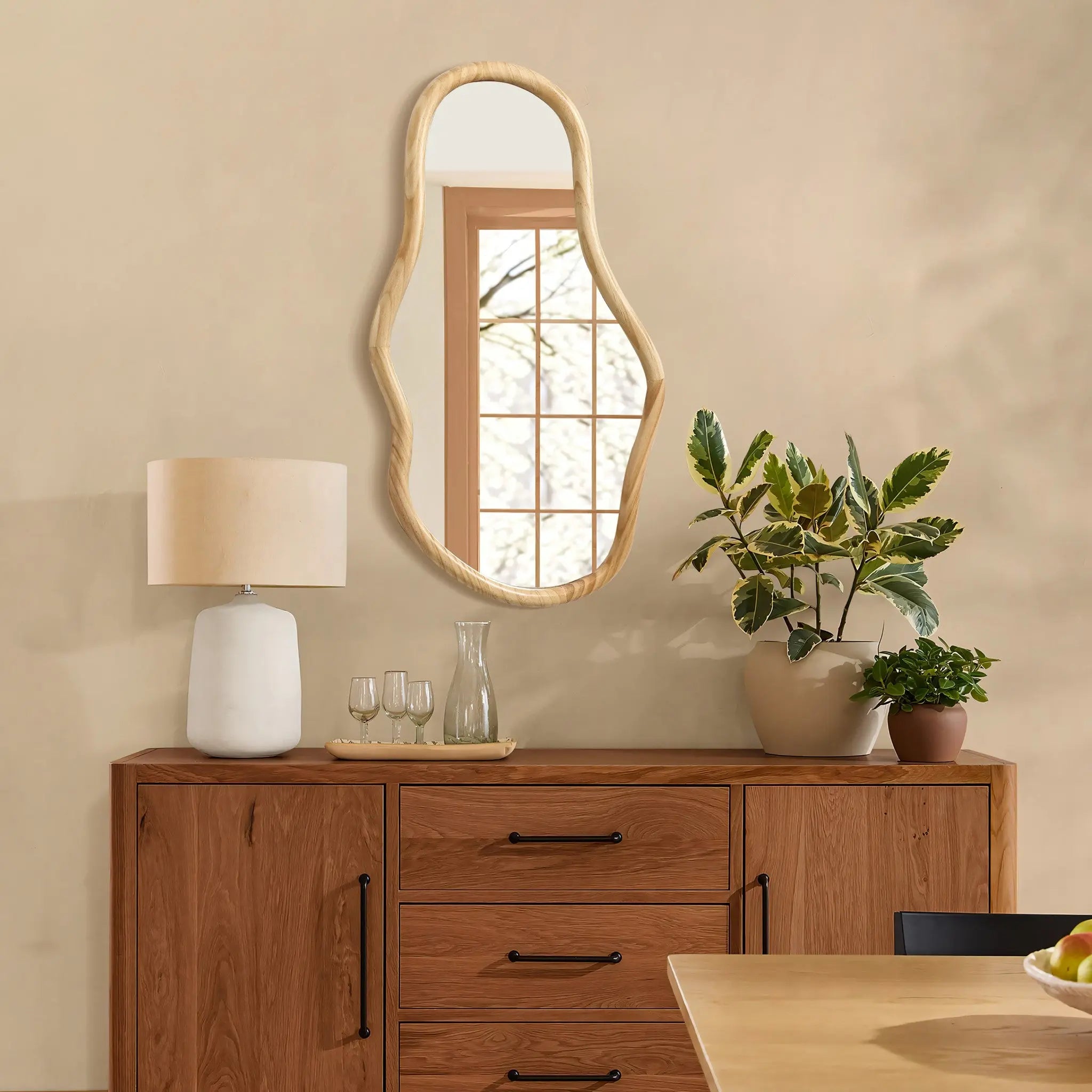 A decorative statement piece, the wooden sideboard crafted from natural pine wood features an irregular oval-shaped mirror, complemented by a lamp and plants. The windows reflection reveals trees outside, enhancing its organic charm.