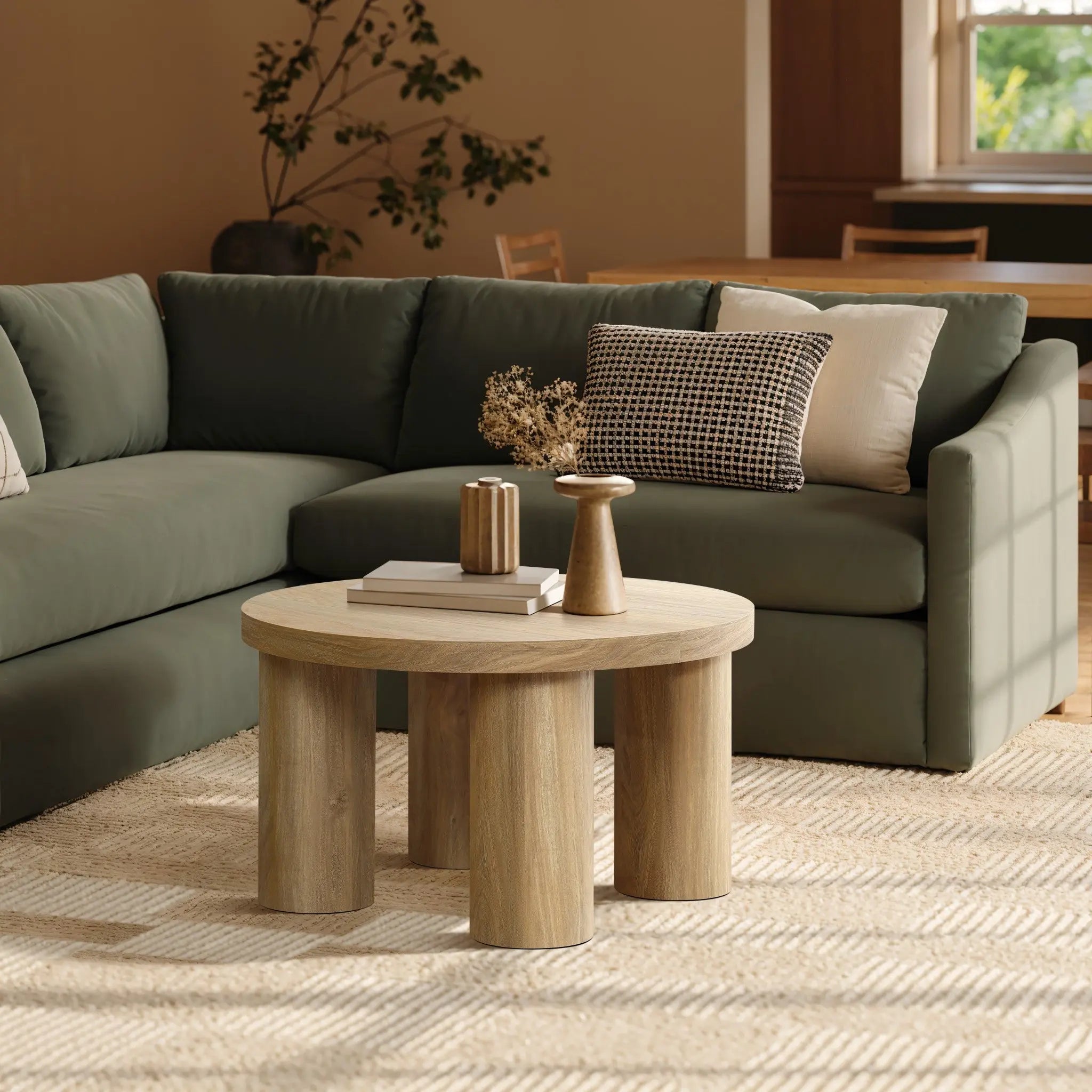 A round oak-finish coffee table with vases is in front of a green sectional sofa. A patterned pillow adorns the sofa. Sunlight illuminates the beige rug, while plants thrive in the background, creating a serene atmosphere.