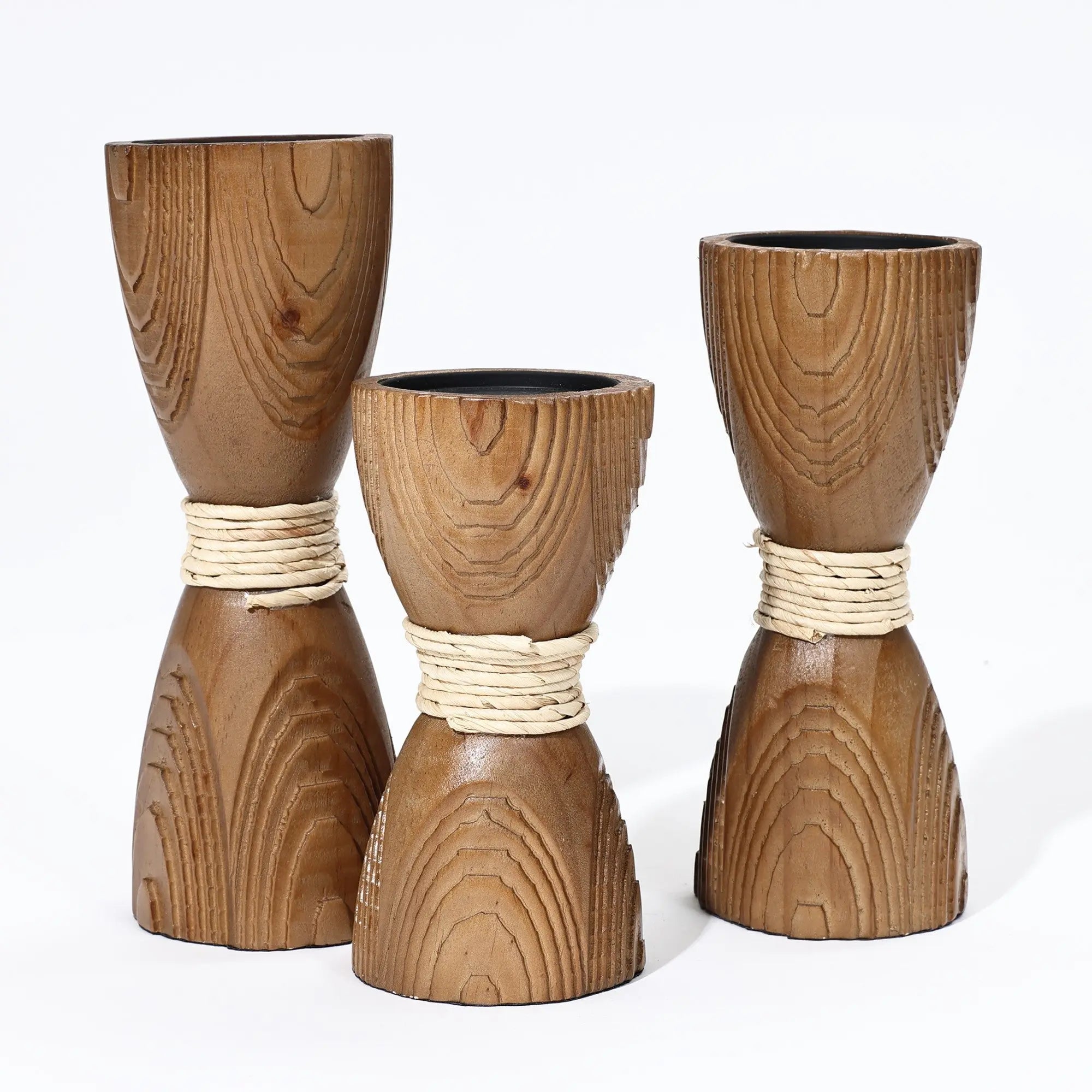 Three wooden hourglass-shaped vases with visible grain patterns, wrapped with white rope in the middle, evoke a handcrafted feel against a plain white background. Their elegant form mimics the allure of candle holders.
