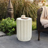 A white, fluted modern side table made of cement with a cup and saucer on top stands next to a wicker chair and a garden gnome statue, adding charm to the outdoor decor of the stone patio.