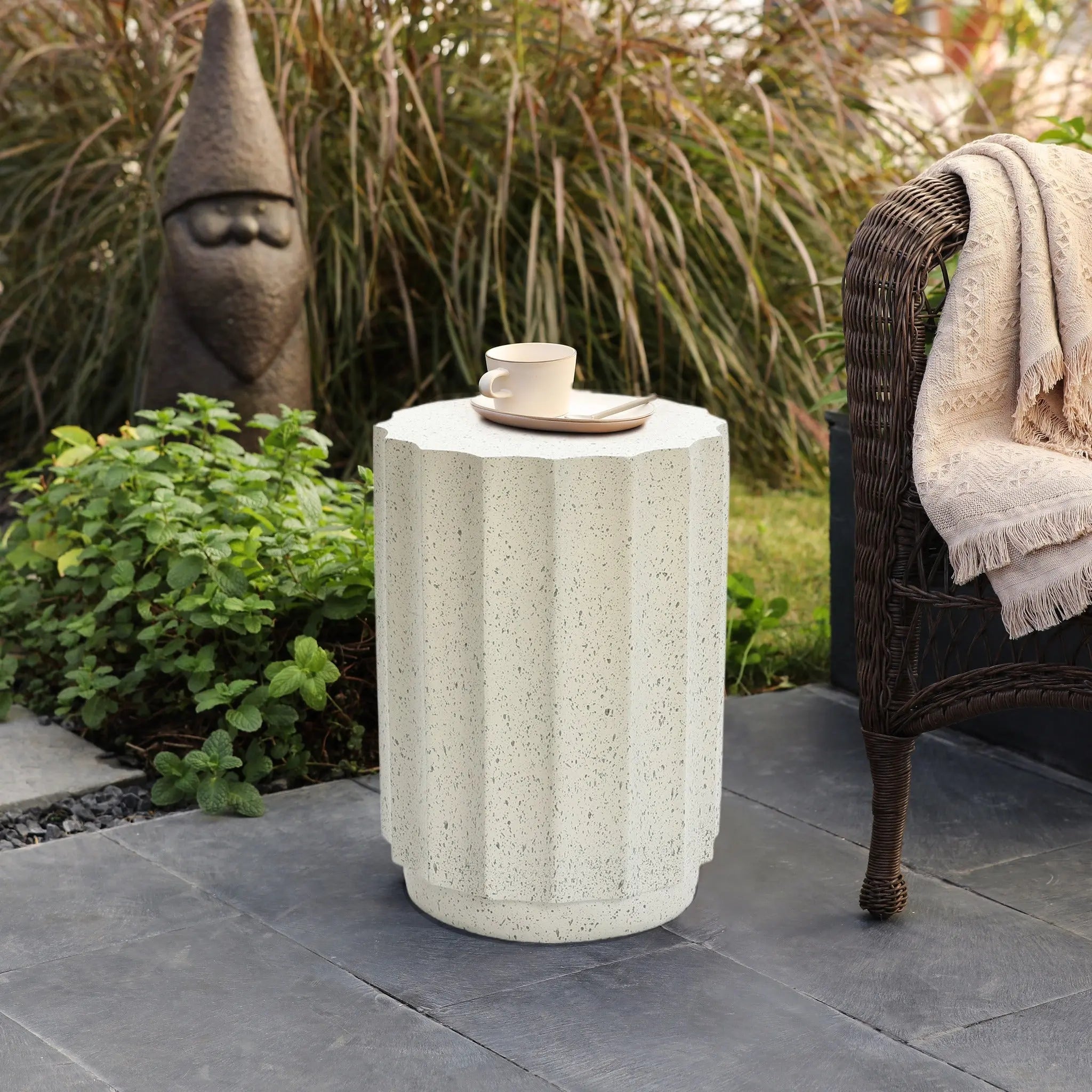 A white, fluted modern side table made of cement with a cup and saucer on top stands next to a wicker chair and a garden gnome statue, adding charm to the outdoor decor of the stone patio.