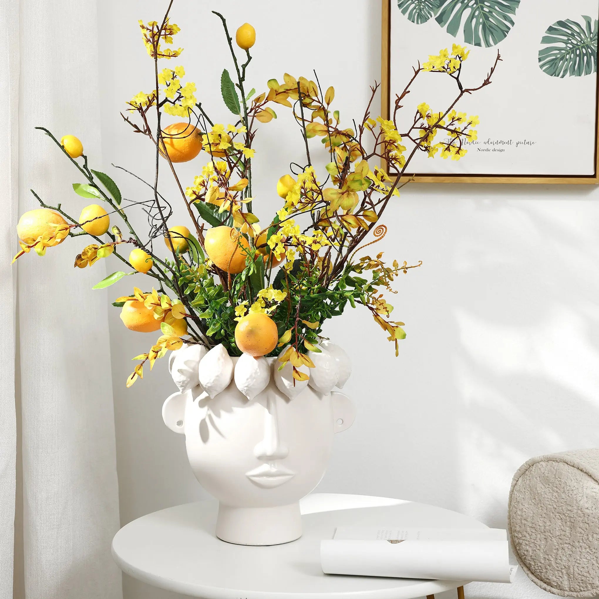A whimsical LuxenHome white ceramic bust head vase, 9.6 high, holds vibrant yellow flowers and lemons on a round table against a white wall. A framed botanical print is partially visible in the background.