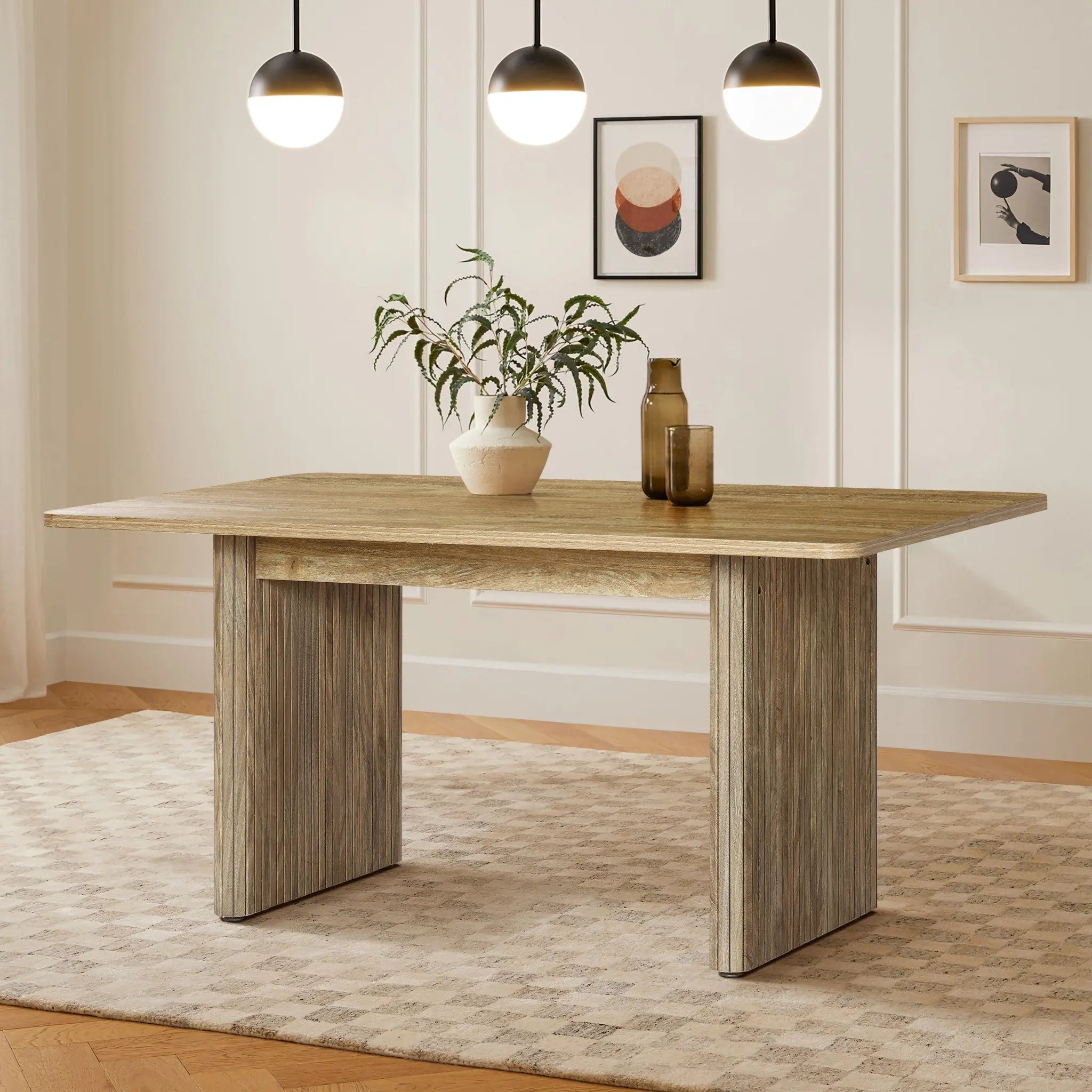 The minimalist room features a LuxenHome 63 wide Aria wood finish dining table for six, set on a patterned rug. The spacious surface holds a potted plant and two bottles. Above, three spherical pendant lights illuminate the space, complemented by framed artwork on the wall.