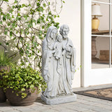 A marble-like polyresin statue depicting a robed family of three graces the patio, surrounded by potted plants near glass doors. This Holy Family statue seamlessly blends into the serene setting, adding an element of religious decor to the peaceful environment.