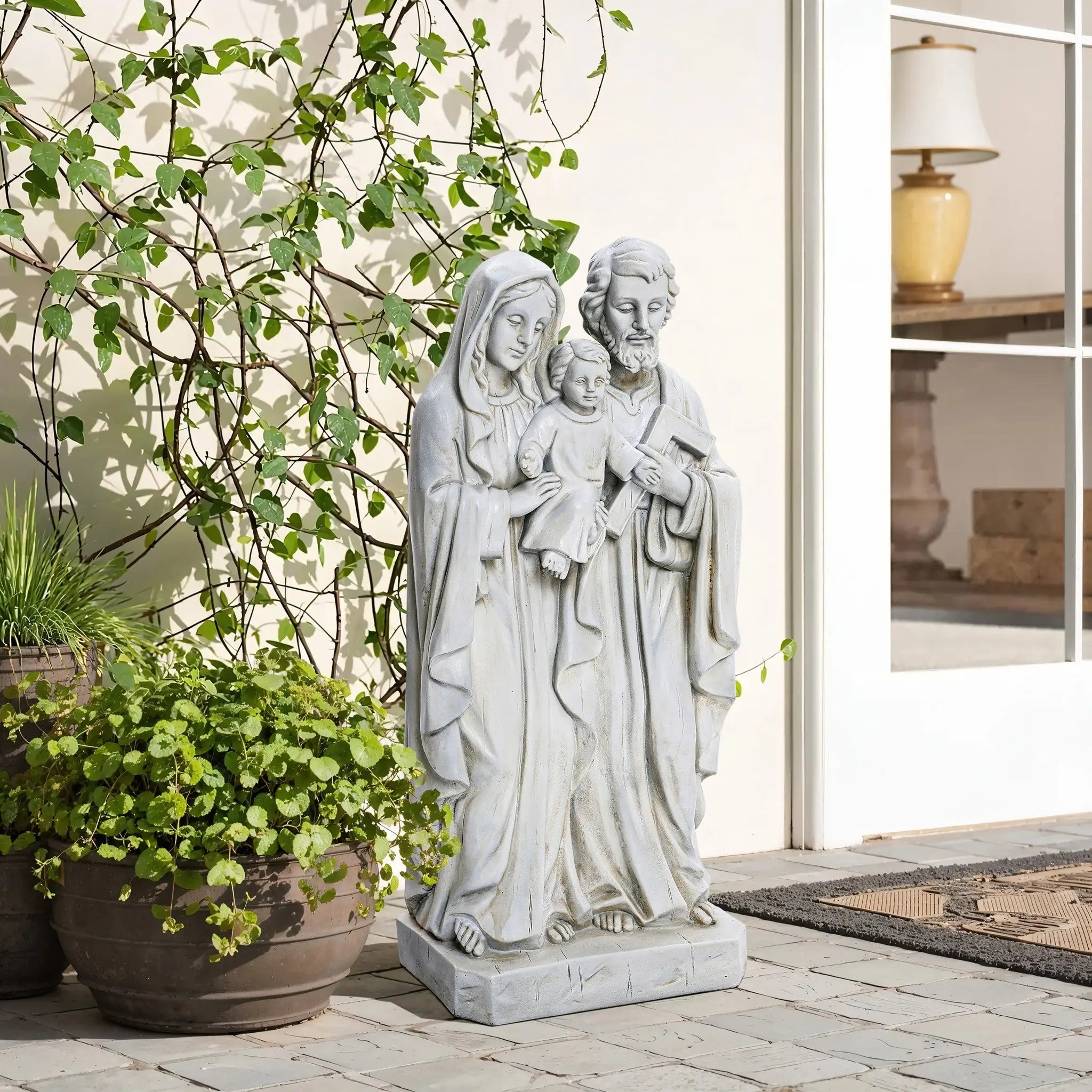 A marble-like polyresin statue depicting a robed family of three graces the patio, surrounded by potted plants near glass doors. This Holy Family statue seamlessly blends into the serene setting, adding an element of religious decor to the peaceful environment.