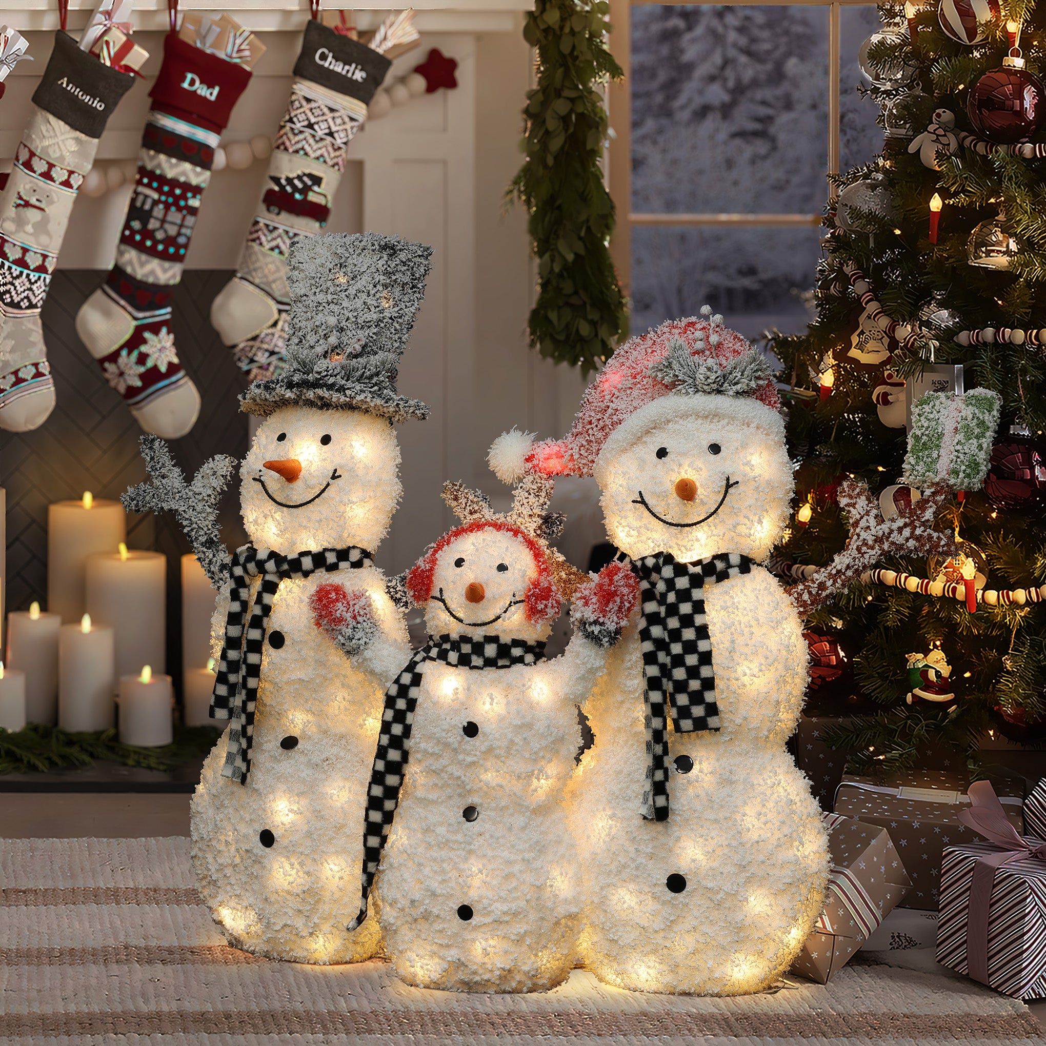 Three lighted snowmen with black and white scarves stand in front of a Christmas tree and fireplace decorated with stockings and candles, creating a charming snowman decoration perfect for your outdoor Christmas decor.