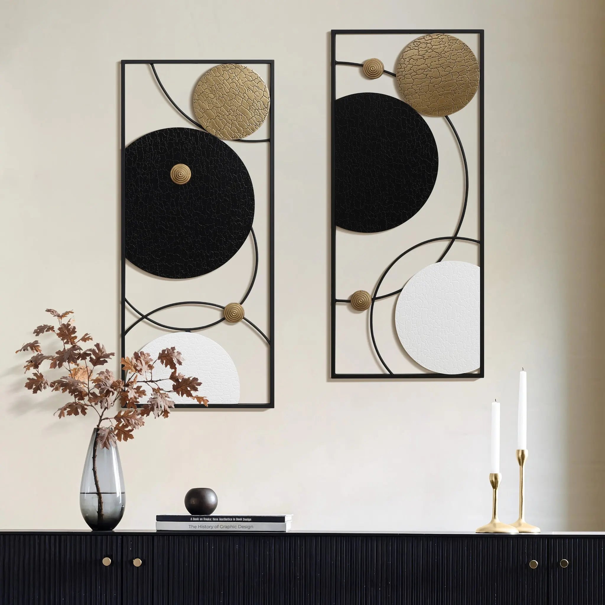 Modern abstract wall decor in a stunning two-piece set showcases circular designs in black, white, and gold. Placed on a sleek black sideboard, the art is complemented by dried flowers in a vase and the soft glow of two lit candles.