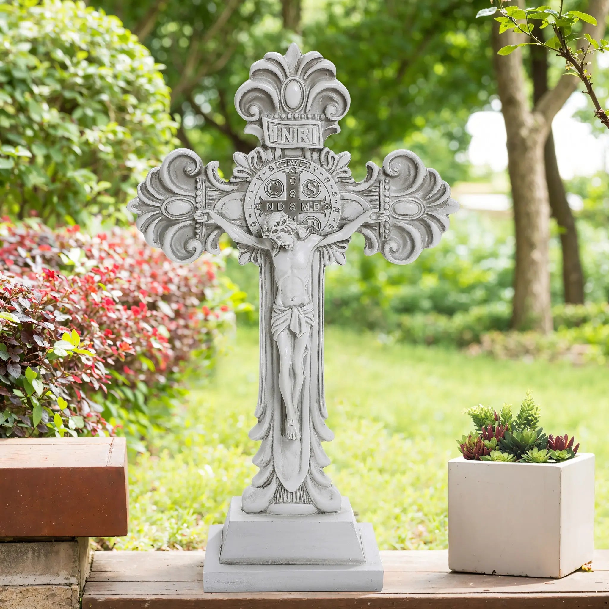 A stone cross sculpture, adorned with ornate designs and a crucifix, stands on a small platform in a garden setting. Surrounded by greenery and red foliage, it embodies the Christian faith and adds a serene touch for religious occasions.