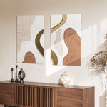 Two modern art pieces with beige, brown, and gold curves hang above a wooden console. The console holds black candlesticks and a spotted vase, creating a chic home decor vibe. Sunlight casts shadows on the brown and white wall art.