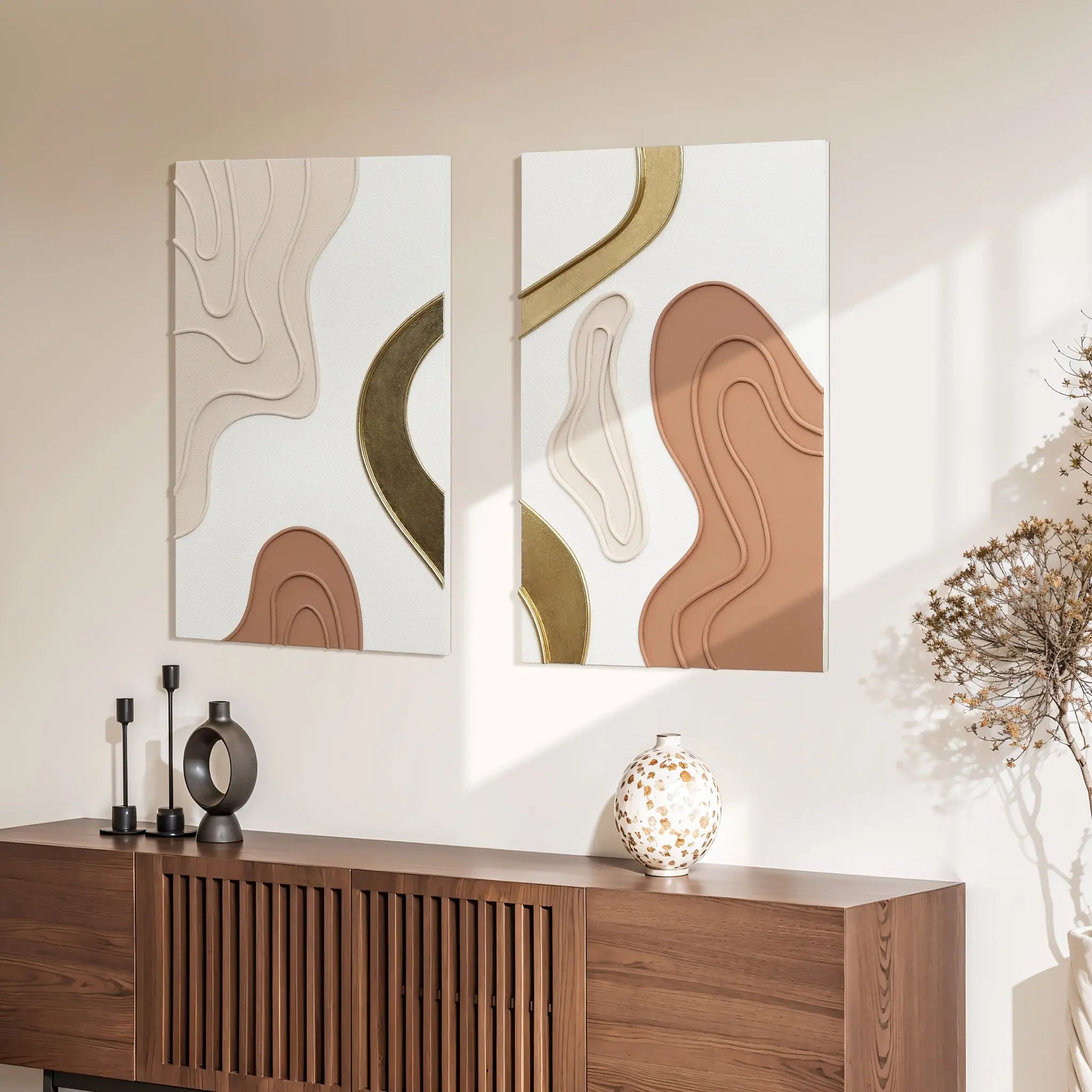 Two modern art pieces with beige, brown, and gold curves hang above a wooden console. The console holds black candlesticks and a spotted vase, creating a chic home decor vibe. Sunlight casts shadows on the brown and white wall art.