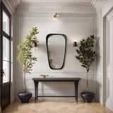 Elegant hallway with a black console table, an elongated mirror for distortion-free viewing, wall sconces, and two potted trees on a herringbone wood floor.