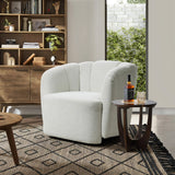 A cozy living room displays an Amber ivory boucle swivel barrel accent chair by LuxenHome, a wooden side table with a bottle and glass, a geometric-patterned rug, a wooden shelf with decorative items, and a large window framing lush greenery.