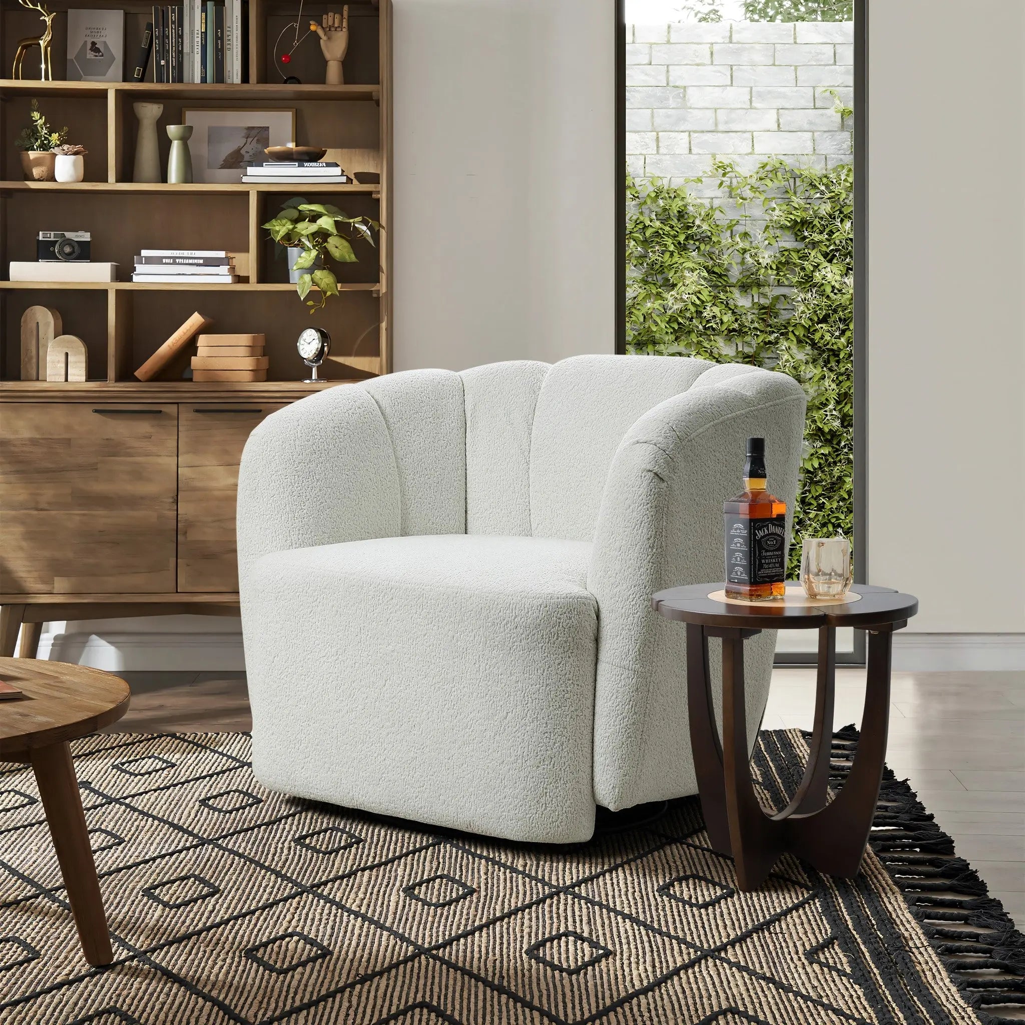 A cozy living room displays an Amber ivory boucle swivel barrel accent chair by LuxenHome, a wooden side table with a bottle and glass, a geometric-patterned rug, a wooden shelf with decorative items, and a large window framing lush greenery.