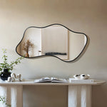 An asymmetrical mirror on a beige wall reflects a simple painting. Below, a white console table embodies modern minimalism, holding a black vase with plants, a book, and a white sculpture.