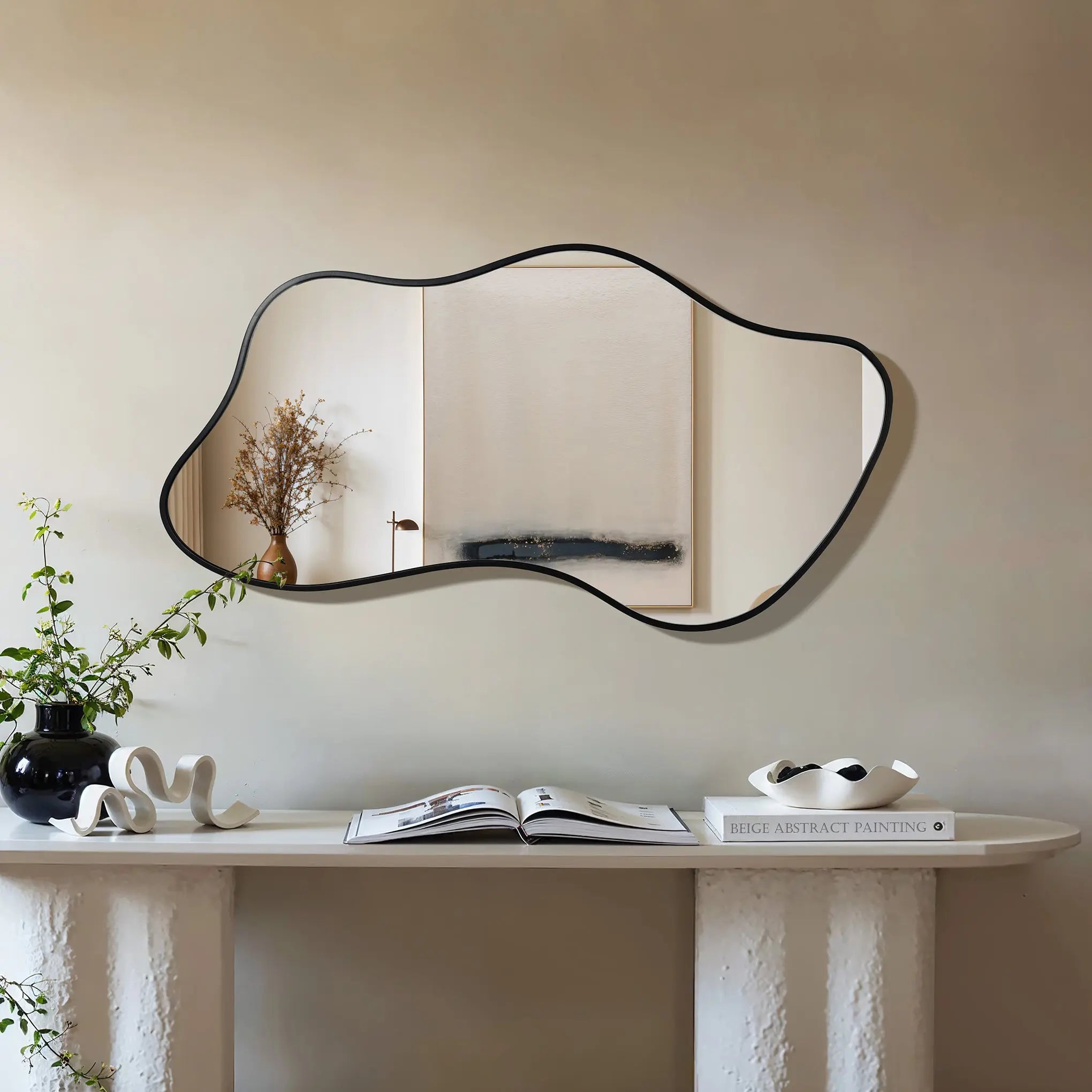 An asymmetrical mirror on a beige wall reflects a simple painting. Below, a white console table embodies modern minimalism, holding a black vase with plants, a book, and a white sculpture.