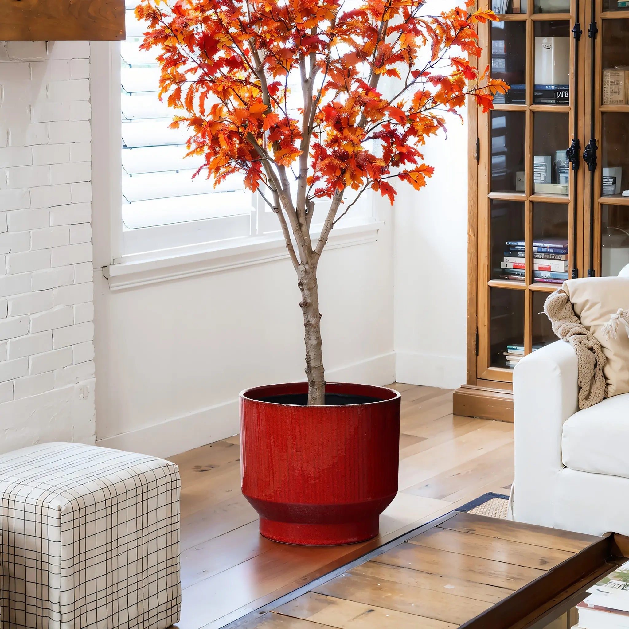 A cozy living room showcases a bright orange artificial maple tree in a LuxenHome burgundy red glazed round ceramic planter, 11.61 h, alongside a white sofa, plaid ottoman, wooden coffee table, and a tall wooden cabinet with glass doors against the white brick wall.