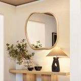 A wooden table features a plant in a glass vase, two black bowls, and a decorative lamp. Above, an irregularly shaped wall mirror with a natural pine frame is mounted on the beige wall, adding charm to the home decoration.