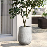 A potted tree thrives in a large, tapered round planter crafted from MgO material, featuring a subtle speckled design. Its perfectly placed on the tiled patio beside a sliding glass door, with a cozy sofa invitingly visible in the background.