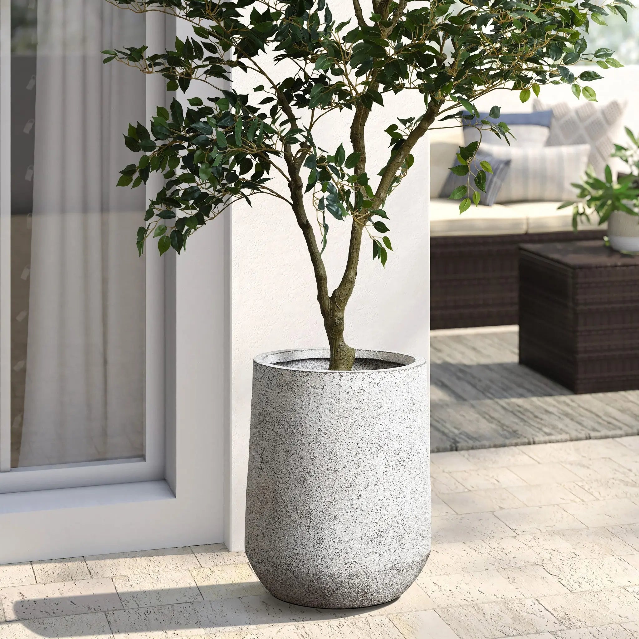 A potted tree thrives in a large, tapered round planter crafted from MgO material, featuring a subtle speckled design. Its perfectly placed on the tiled patio beside a sliding glass door, with a cozy sofa invitingly visible in the background.