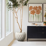 A tall plant in a White and Beige Geometric Patterned Round Planter from LuxenHome is beside a black sideboard with a white top. Framed botanical prints hang above. Large windows on the left flood natural light into the modern, minimalist room, accentuating its geometric design elements.