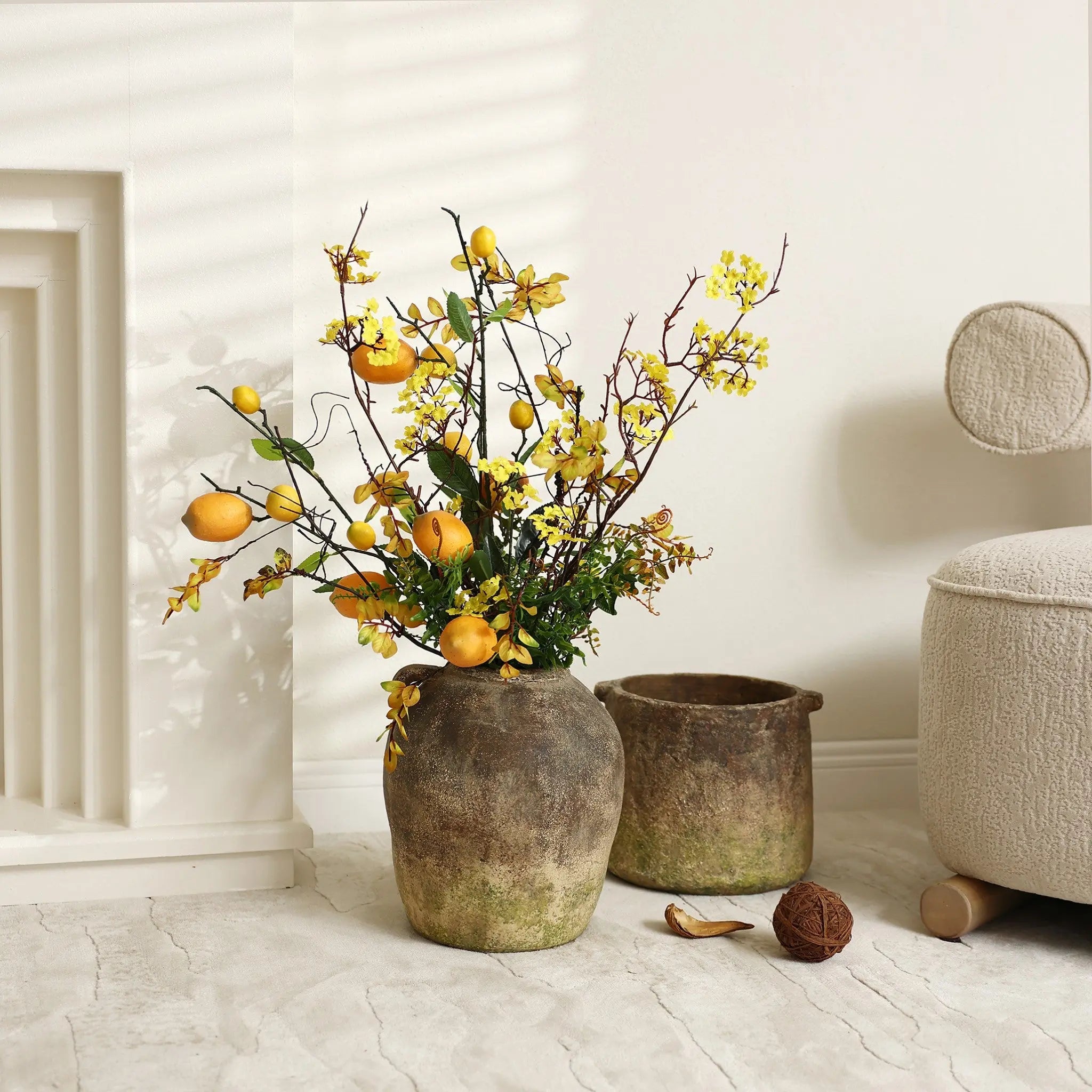 A rustic terracotta vase with a handcrafted weathered finish holds yellow flowers and lemons next to an empty pot and a ball of twine on a light-colored floor.