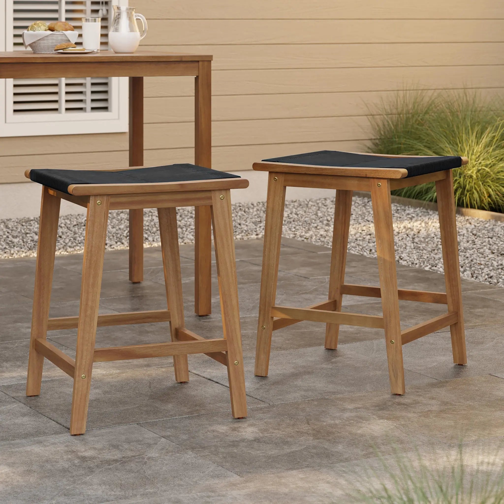 Two stylish Acacia wood barstools with black rope-woven seats grace a stone patio next to a wooden table. A beige building and lush greenery provide a serene backdrop.