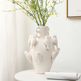 A LuxenHome 14.4 white ceramic birds vase sits on a round white table, exuding elegance with branches of small green leaves. The background, with a white wall and framed picture, epitomizes modern home decor.