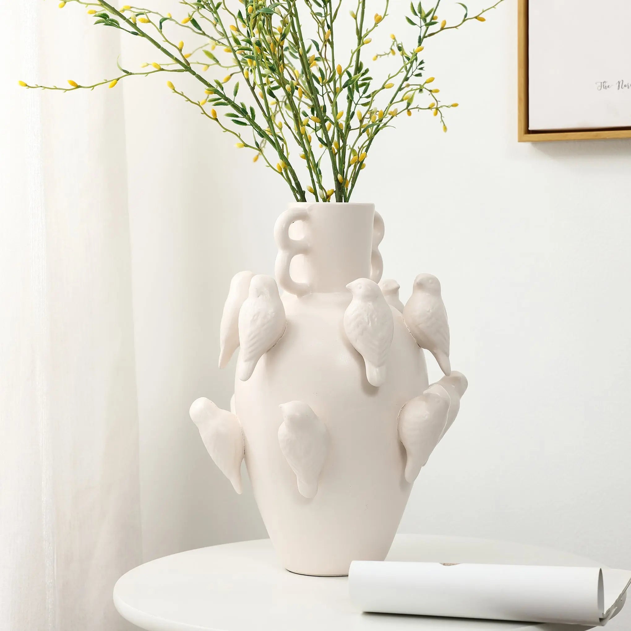 A LuxenHome 14.4 white ceramic birds vase sits on a round white table, exuding elegance with branches of small green leaves. The background, with a white wall and framed picture, epitomizes modern home decor.