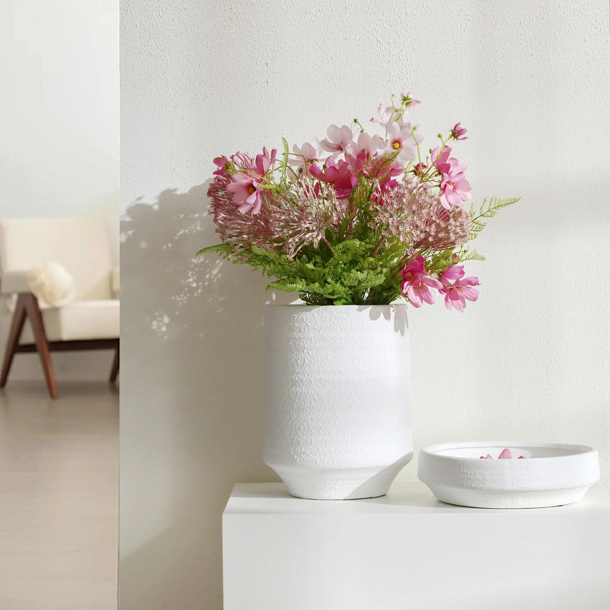 A 12-inch tall white ceramic vase with pink flowers and green leaves sits gracefully on a white surface near a white wall, complemented by a matching white dish. This versatile piece effortlessly enhances both indoor and outdoor spaces.