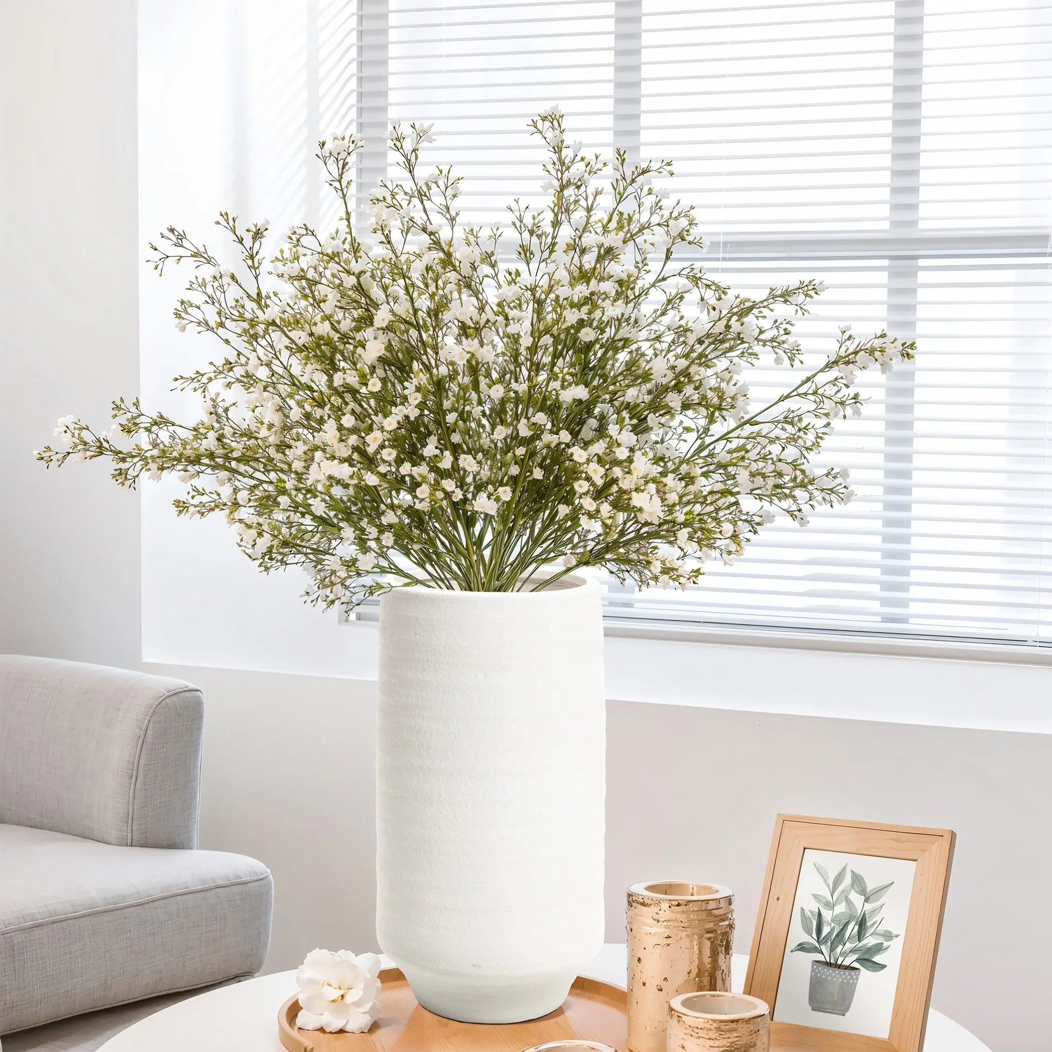 A white terracotta vase with white flowers graces the table, alongside two candles and a petite framed botanical print, in a room featuring a gray couch and window blinds.
