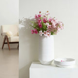 A white terracotta vase with pink and white flowers graces a white table beside an empty bowl. In the background, a beige chair adds warmth to this serene indoor scene.